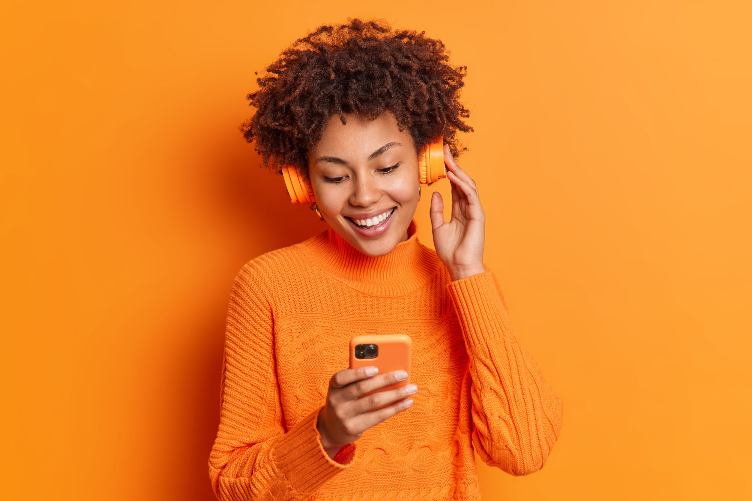 portrait-cheerful-woman-chooses-song-listen-from-her-playlist-enjoys-good-sound-headphones-concentrated-smartphone-display-dressed-casually-isolated-orange-wall