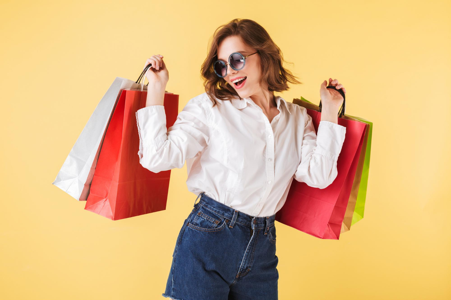 portrait-happy-lady-sunglasses-standing-with-colorful-shopping-bags-hands-pink-background-young-woman-standing-white-shirt-denim-shorts