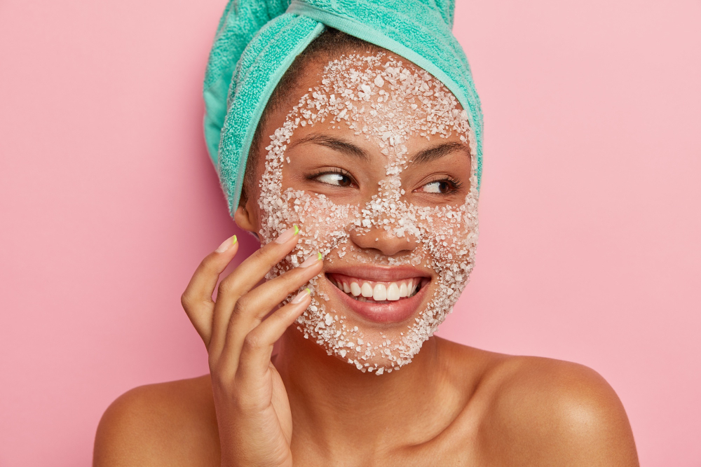 close-up-portrait-glad-woman-massages-cheeks-applies-sea-salt-scrub-looks-away-has-gentle-smile-shows-white-teeth-wears-turquoise-towel-happy-get-spa-treatments